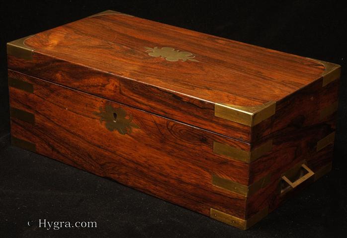 Figured rosewood writing box with brass corners,  straps, and  countersunk carrying handles having a side drawer and canceled secret drawers. The writing surface is framed by a cross -banding of rosewood and has been recovered with an embossed leather skiver.  The escutcheons both in front of the lock and the top are of ornate shape with spear like  fleur dis lis motif  to the side  The box has a working lock and key. Circa 1840.  Enlarge Picture